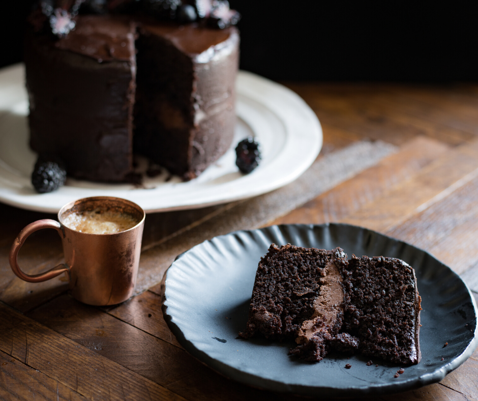 slice of chocolate cake on plate, next to coffee cup and whole cake on a separate plate