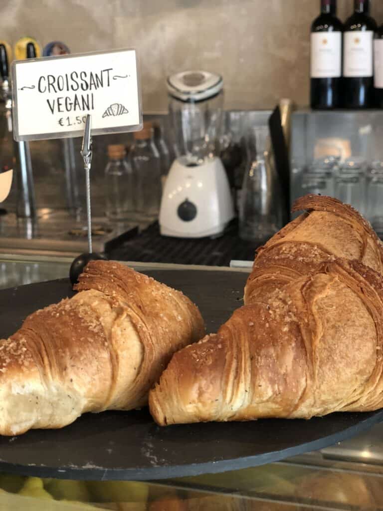 Two large vegan croissants displayed on a round black tray, labeled CROISSANT VEGANI with a price of €1.50. A blender and wine bottles are visible in the background, evoking a charming Italian café ambiance.