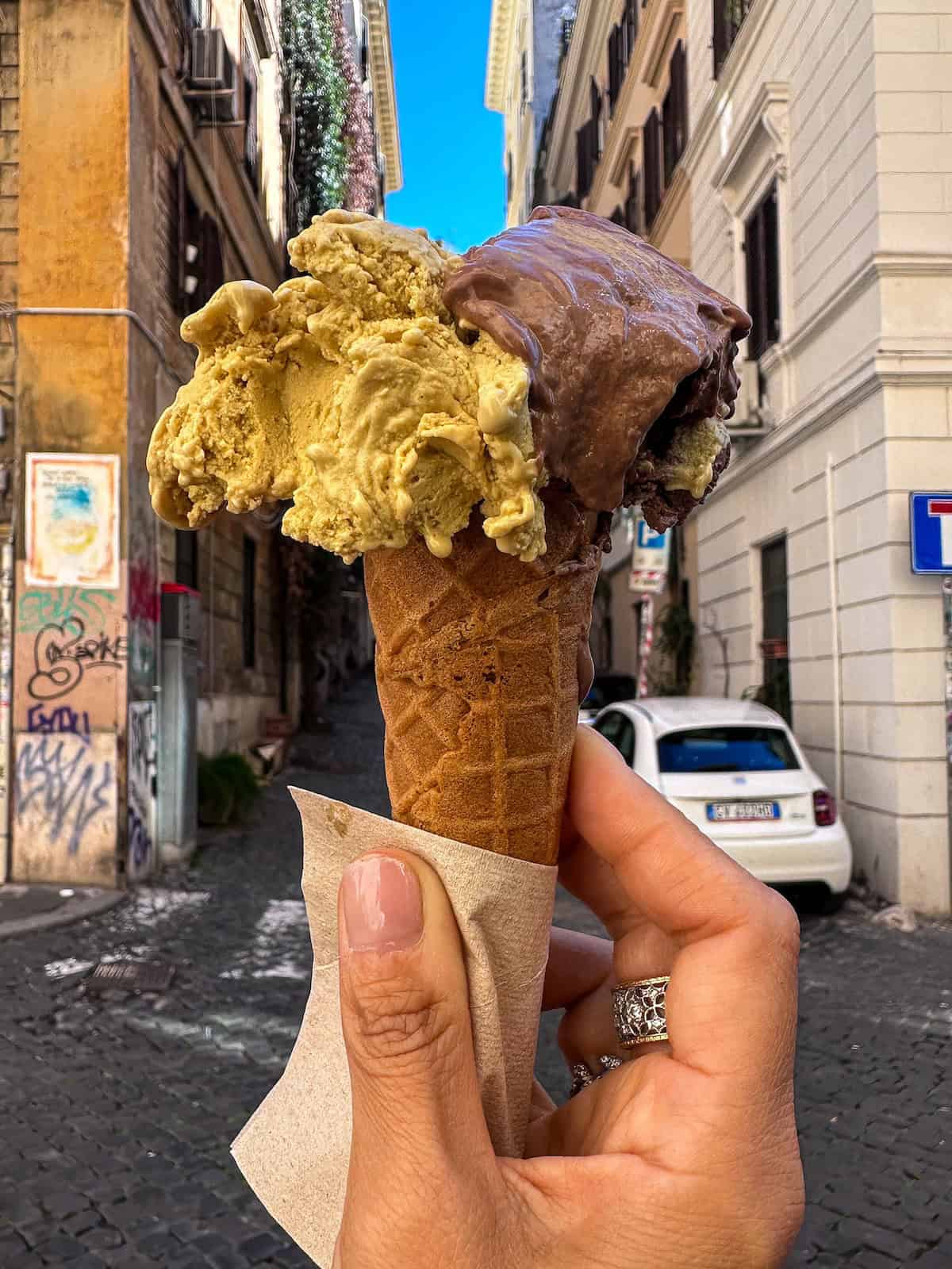 A person holds a waffle cone with two scoops of gelato—pistachio and chocolate—against a narrow, quaint Roman street in Italy, with cobblestones, parked cars, and buildings adorned with street art and ivy-covered walls.