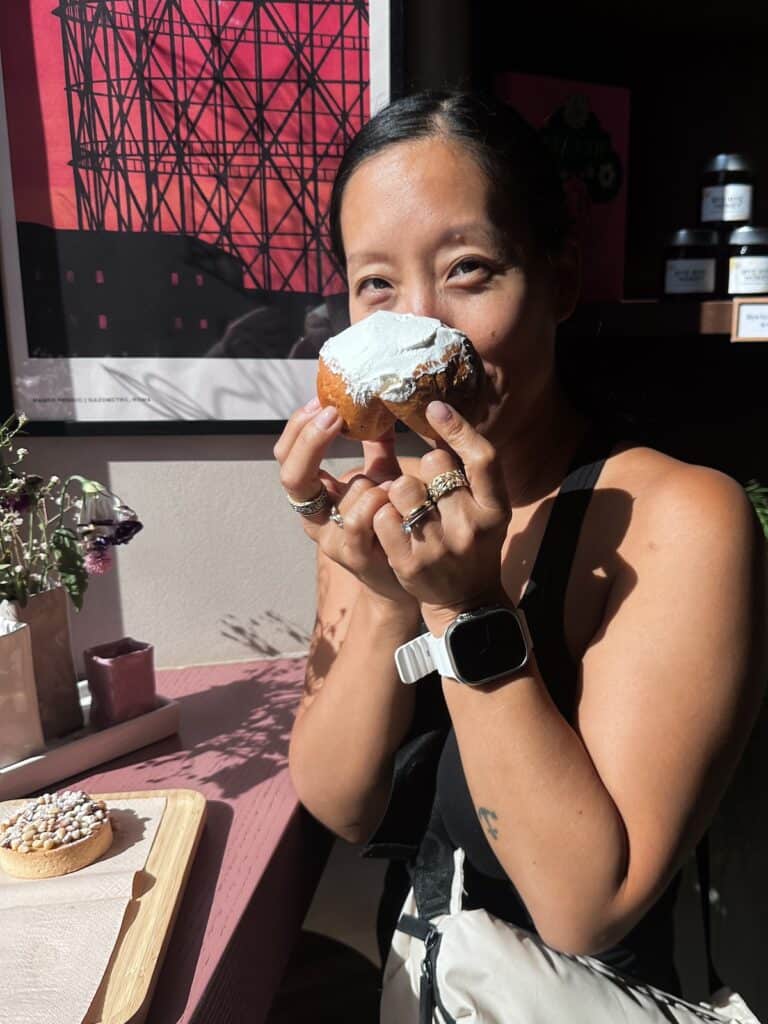 Joanne holding a frosted vegan pastry near her face, smiling. She is in a room with an Italian flair, featuring a pink table, a poster, and sunlight streaming through the window. Another sprinkle-adorned pastry rests nearby on the table.