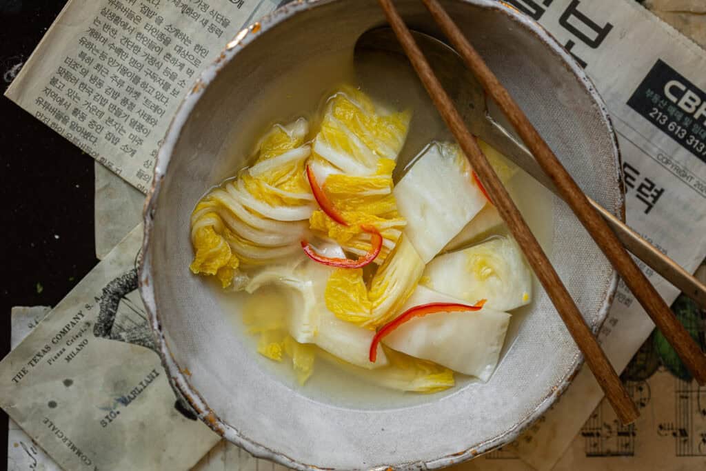 A bowl of white kimchi with slices of red chili peppers in a light-colored liquid. There are wooden chopsticks on the side, and the bowl is placed on sheets of old newspaper.