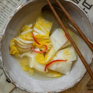 A bowl of white kimchi or Baek-Kimchi soup with slices of red chili sits on a table covered with Korean newspaper pages. Brown wooden chopsticks rest on the edge, whispering tales of moms recipe in every bite.