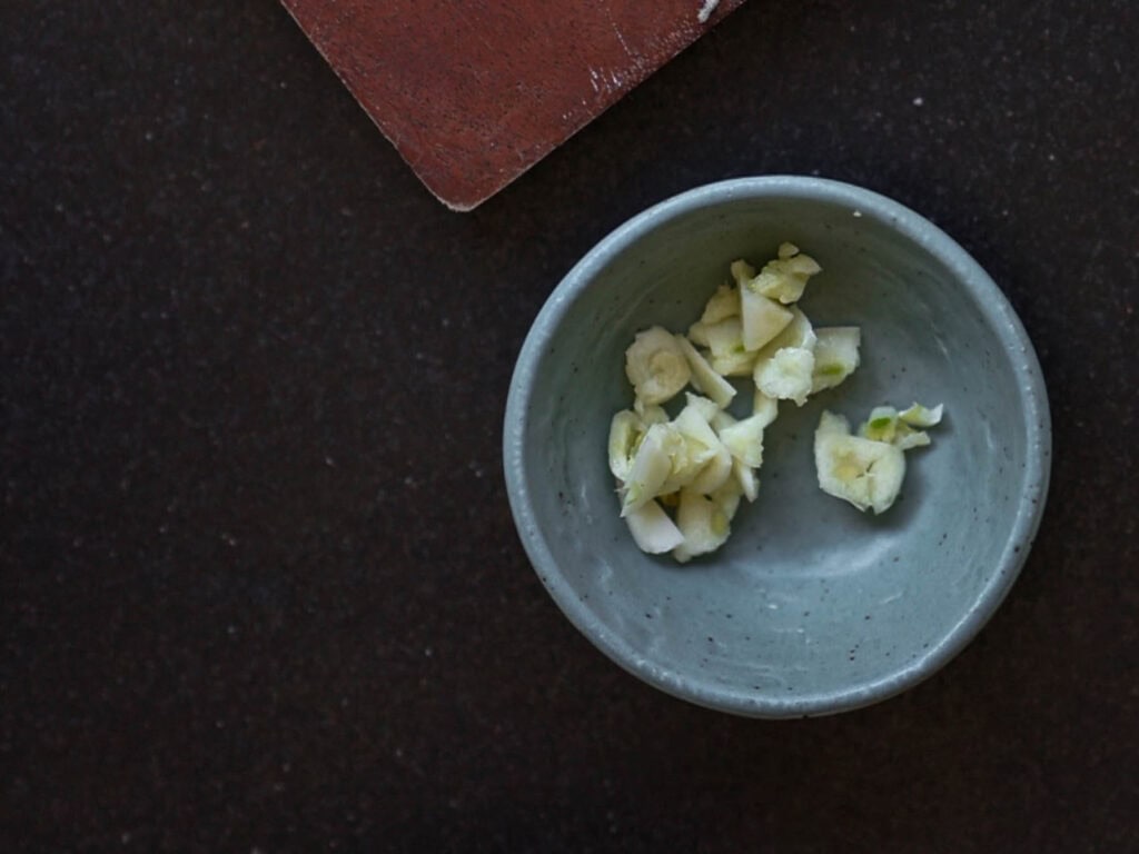 A small green bowl filled with chopped garlic pieces is placed on a dark countertop. A wooden board is partially visible in the top left corner.