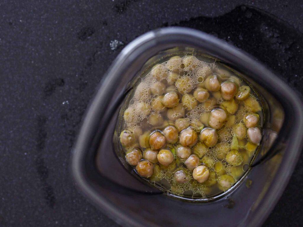 A top view of a blender container filled with soaked chickpeas and their cooking liquid. The chickpeas are partially visible through the transparent sides, with bubbles forming on the surface of the liquid. The container is on a dark countertop.