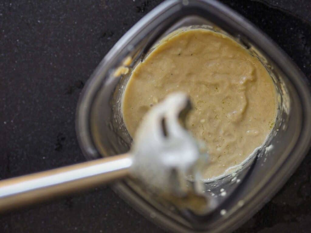 Close-up of a blending container with a creamy light brown mixture inside. An immersion blender, partially submerged, is positioned in the center. The container rests on a dark countertop.