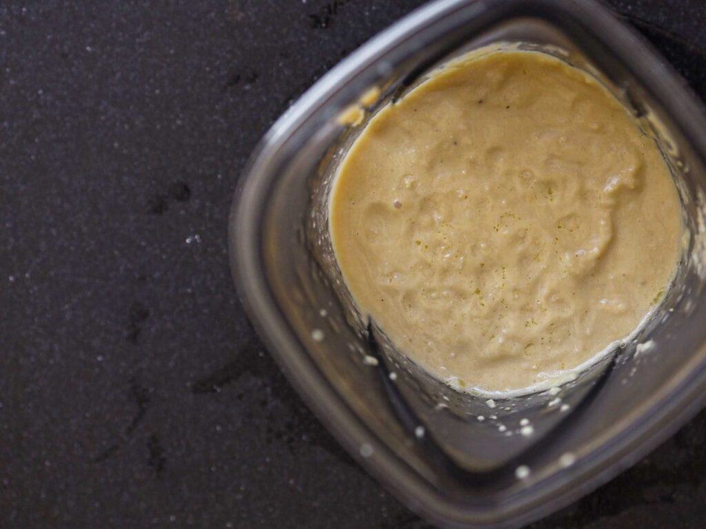 A top-down view of a blender containing smooth, creamy chickpea sauce. The background is a dark, textured surface, highlighting the contrasting pale color of the hummus inside the blender.