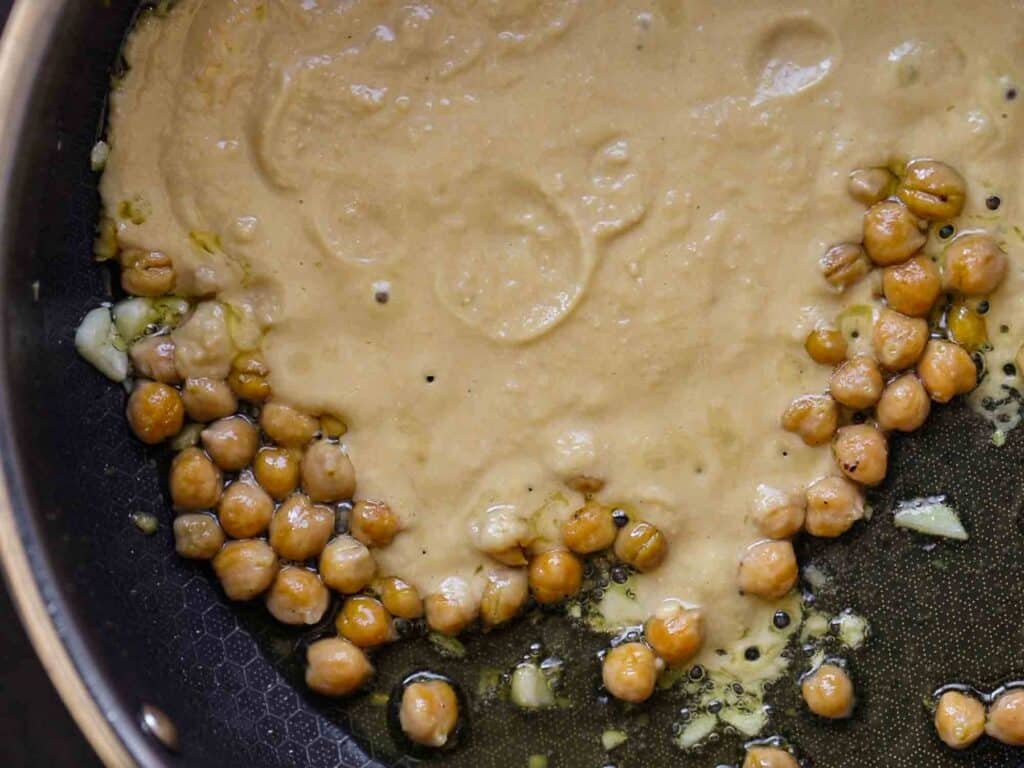 Chickpeas and a creamy sauce cooking in a black skillet with bits of minced garlic. The chickpeas are partially covered by the sauce, which is light brown and smooth in texture.