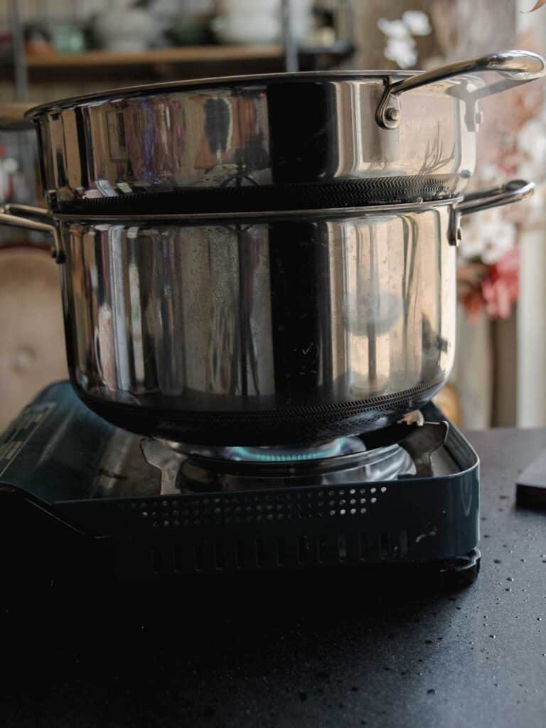 Two stacked stainless steel pots are placed on a portable gas burner. The burner flame is visible, and the background shows blurred kitchen items on shelves.