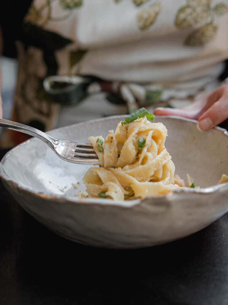 A person twirls fettuccine pasta with a fork in a textured ceramic bowl. The pasta is garnished with fresh green herbs and grated cheese. The background shows the persons torso wearing an apron.