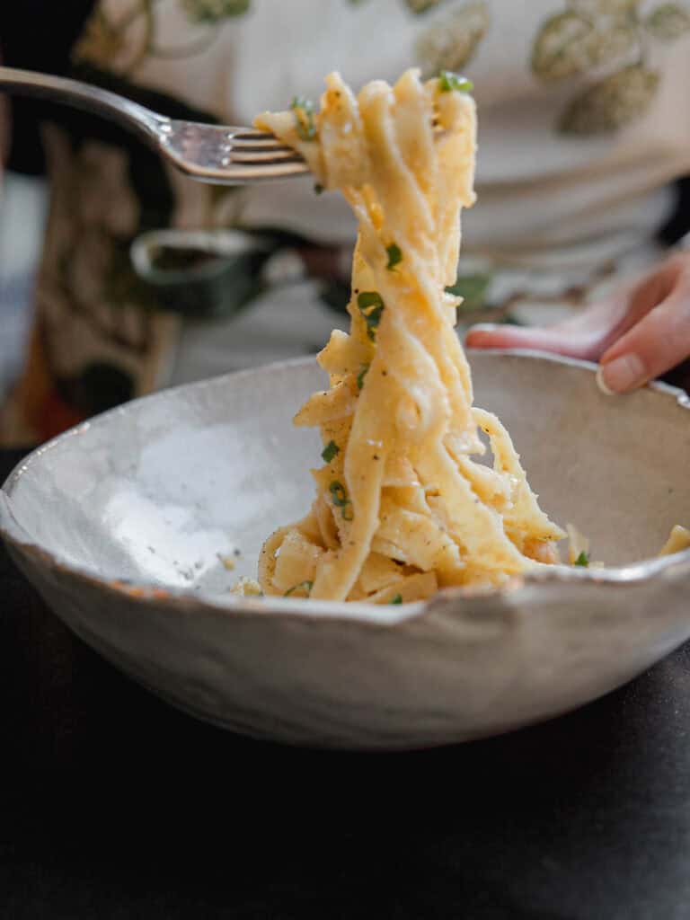 A person twirls a forkful of creamy pasta topped with chopped herbs in a textured, rustic bowl. The background features a person wearing an apron with leafy patterns.