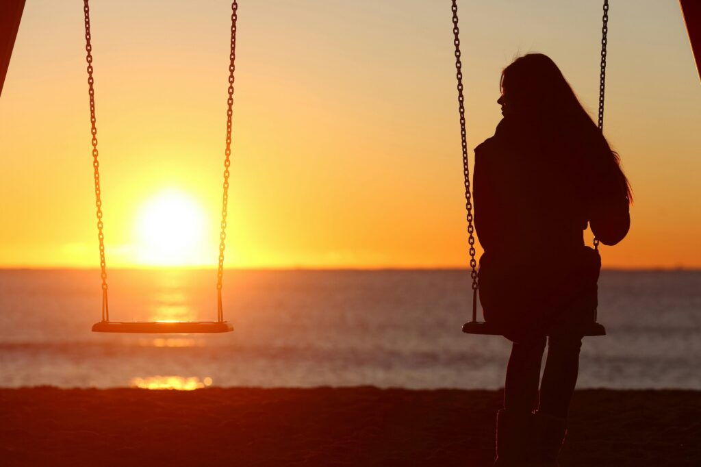 Silhouette of a person sitting on a swing, contemplating friendship as they face the ocean at sunset. The sun is low on the horizon, casting a warm glow over water and sand, while the sky transitions in vibrant hues of orange and yellow. A second swing remains empty.