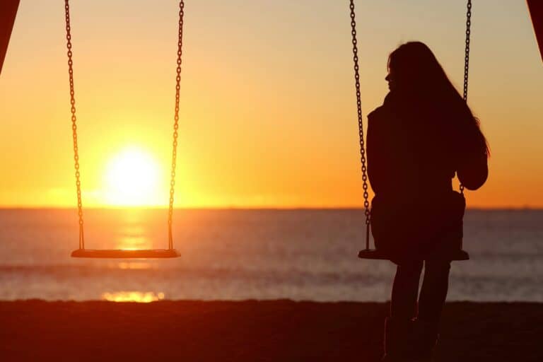 Silhouette of a person sitting on a swing, contemplating friendship as they face the ocean at sunset. The sun is low on the horizon, casting a warm glow over water and sand, while the sky transitions in vibrant hues of orange and yellow. A second swing remains empty.