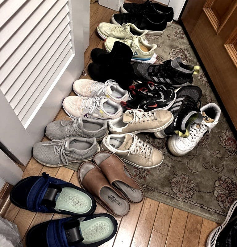 A cluttered pile of various shoes, including sneakers, sandals, and slippers, is scattered on a patterned rug near a wooden door. The scene hints at a family gathering for Thanksgiving.