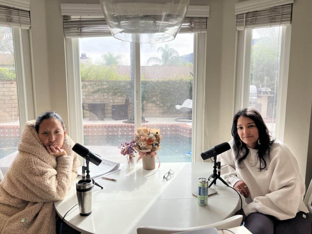 Joanne and Nabiha at a round kitchen table with microphones, recording a podcast. In the cozy room, large windows reveal a pool in the backyard. A vase of flowers and some beverages are on the table.