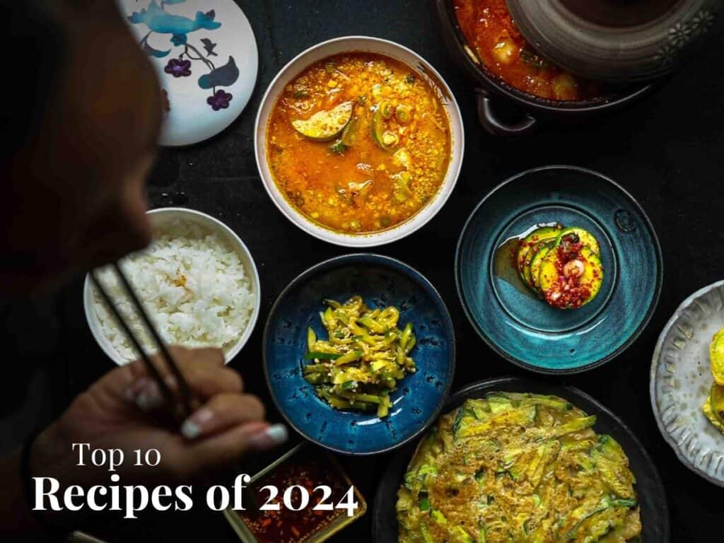 A person eating with chopsticks at a table featuring various dishes, including rice, soup, kimchi, and a vegetable pancake. The image is titled Top 10 Recipes of 2024.