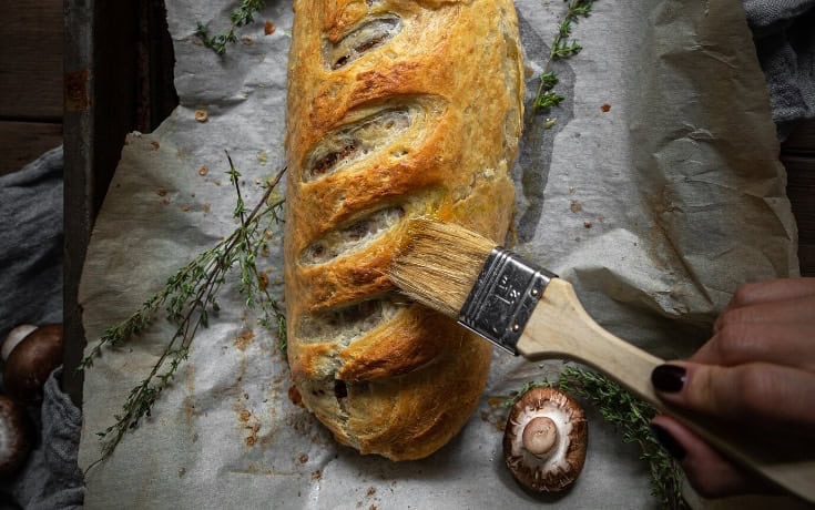 A person brushes a golden-brown Vegan Wellington with glaze on a parchment-lined tray. Fresh thyme sprigs are scattered around, and a mushroom cap is nearby.