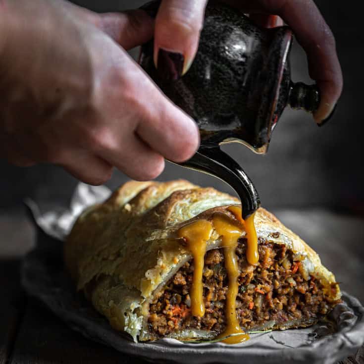 A person pours gravy from a small black jug onto a slice of puff pastry filled with a savory plant-based Italian sausage and vegetable mixture. The golden-brown pastry is on a piece of parchment paper, and the background is softly blurred.