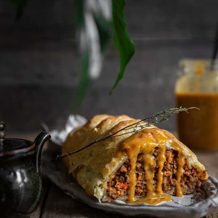 A savory pastry stuffed with plant-based Italian sausage and vegetables, sliced to reveal its filling. Its topped with a drizzle of sauce. A sprig of herbs decorates the pastry. Nearby, theres a small pot and a jar of sauce on a rustic wooden table.