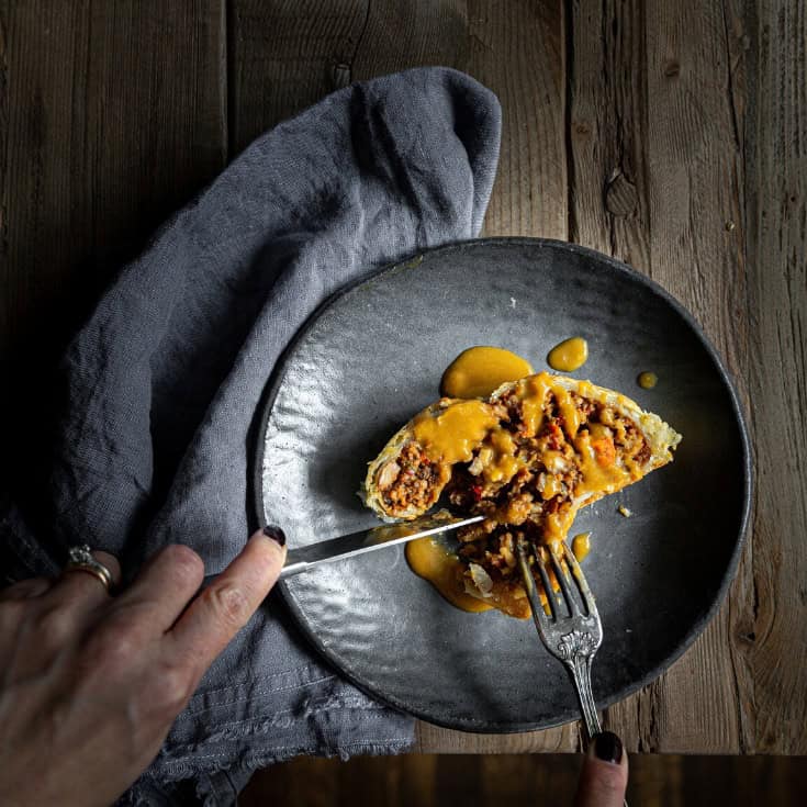 A person cuts into a slice of this Vegan Wellington, which is on a rustic gray plate. The pastry is topped with gravy. A gray cloth is placed beside the plate on a wooden table.