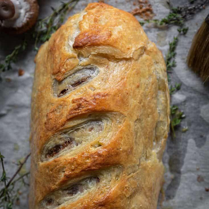 Close-up of a golden-brown loaf of baked pastry with slits on top, resting on parchment paper. Fresh thyme sprigs and a brush are visible in the background, suggesting a savory preparation.