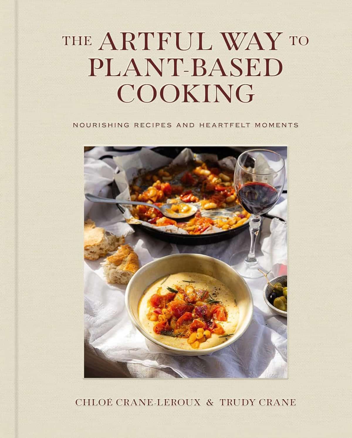 The cover of "The Artful Way to Plant-Based Cooking" serves as a perfect holiday gift, featuring a table adorned with a bowl of stew, a glass of red wine, bread, and a skillet of vegetables. This guide is filled with nourishing recipes and heartfelt moments by Chloe Crane-Leroux & Trudy Crane.