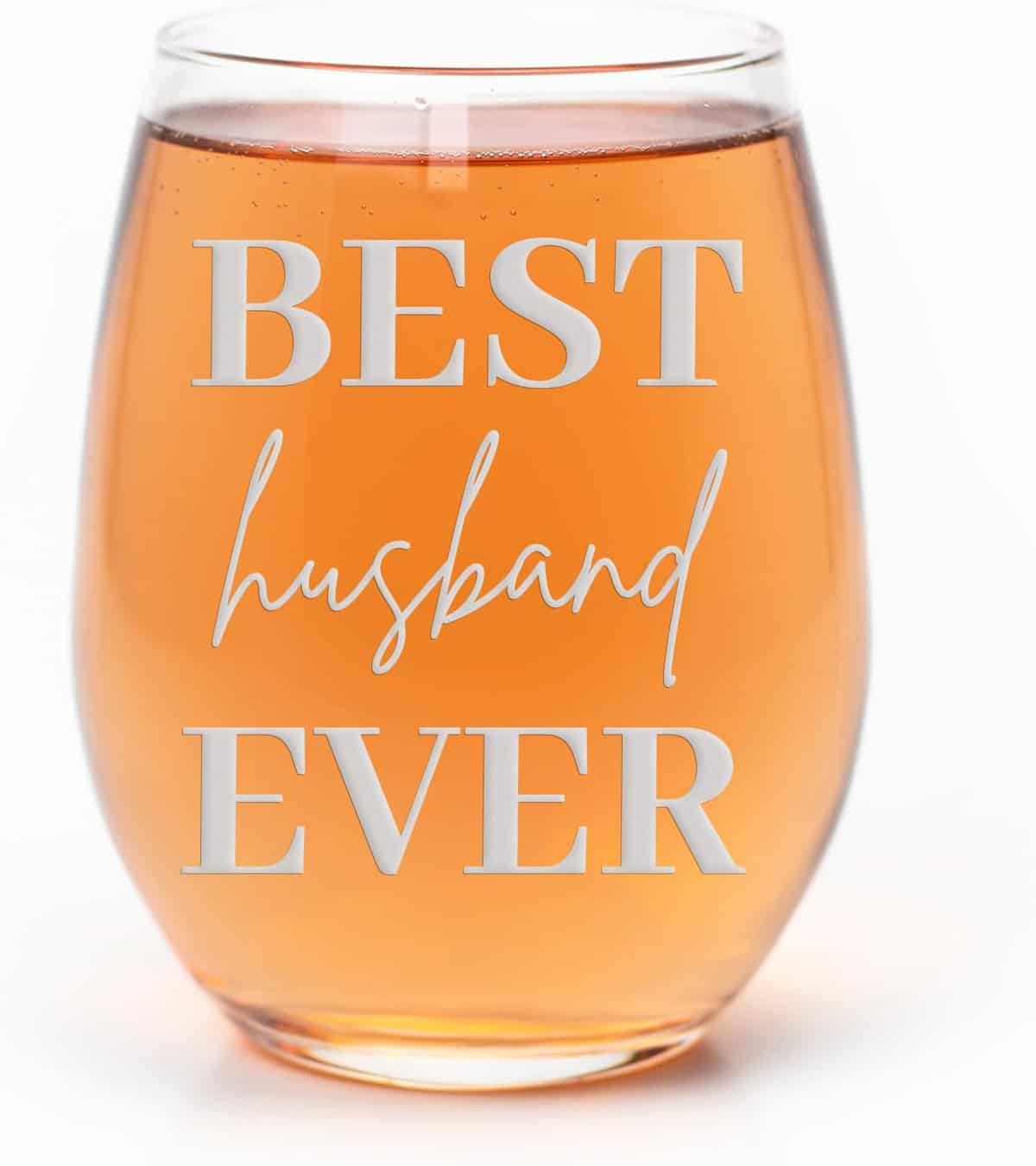 A glass brimming with peach-colored liquid features "Best husband ever" in bold, cursive white letters. Ideal as a holiday gift, this unique item adds a festive charm to any setting. Its simplicity is spotlighted against the white background, making it a standout choice for your 2024 guide.