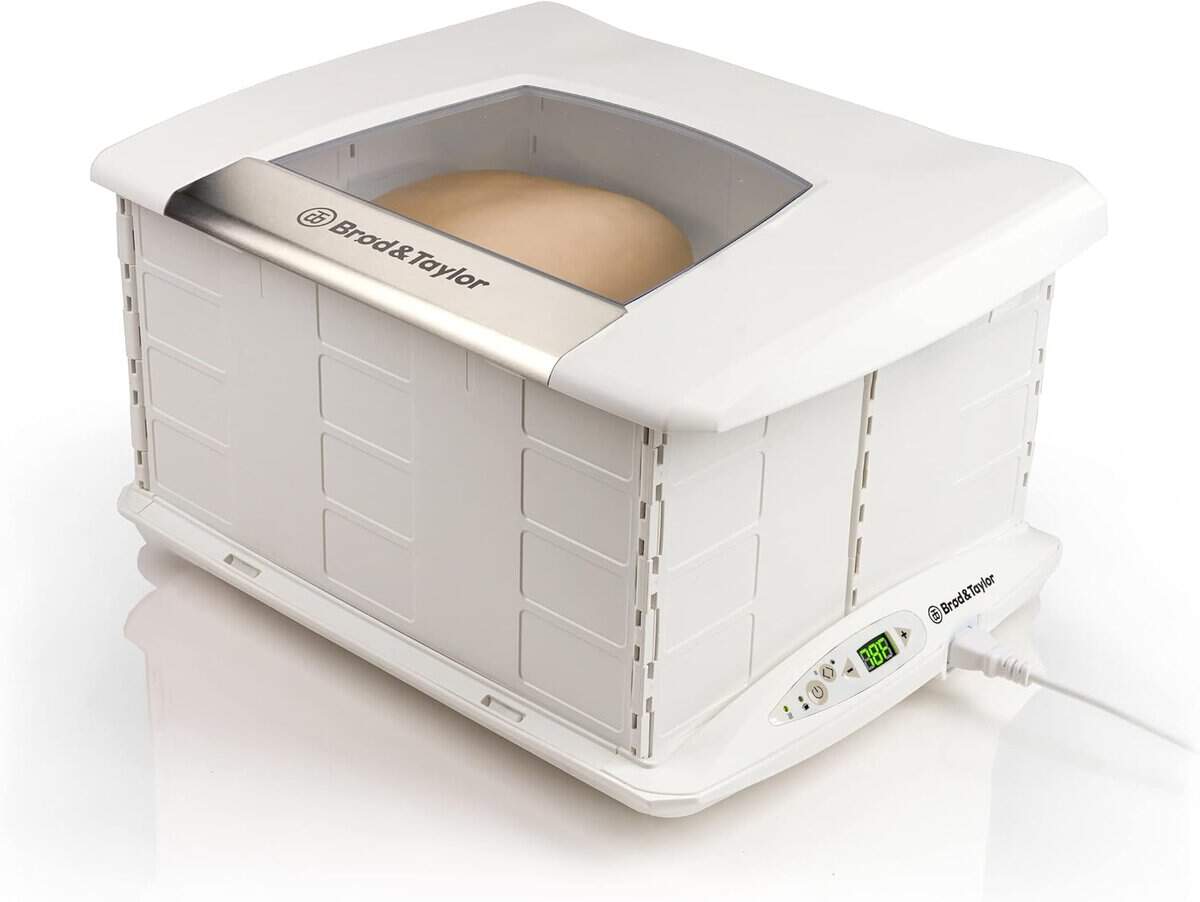 A white bread proofer with dough inside makes a perfect holiday gift, featuring a transparent top window and digital temperature controls on the side, plugged into a power source. The exterior design has vent-like patterns that add an elegant touch to any kitchen countertop.