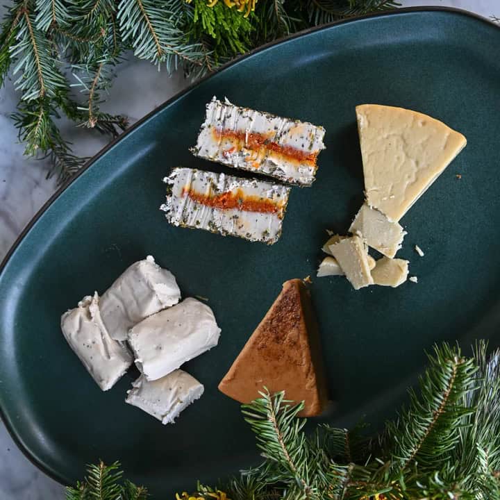 A dark green platter featuring assorted cheese selections, including wedges and slices with visible herbs and spices. Perfect for 2024, the platter is surrounded by green pine branches, enhancing its festive presentation—ideal for a holiday gift guide.