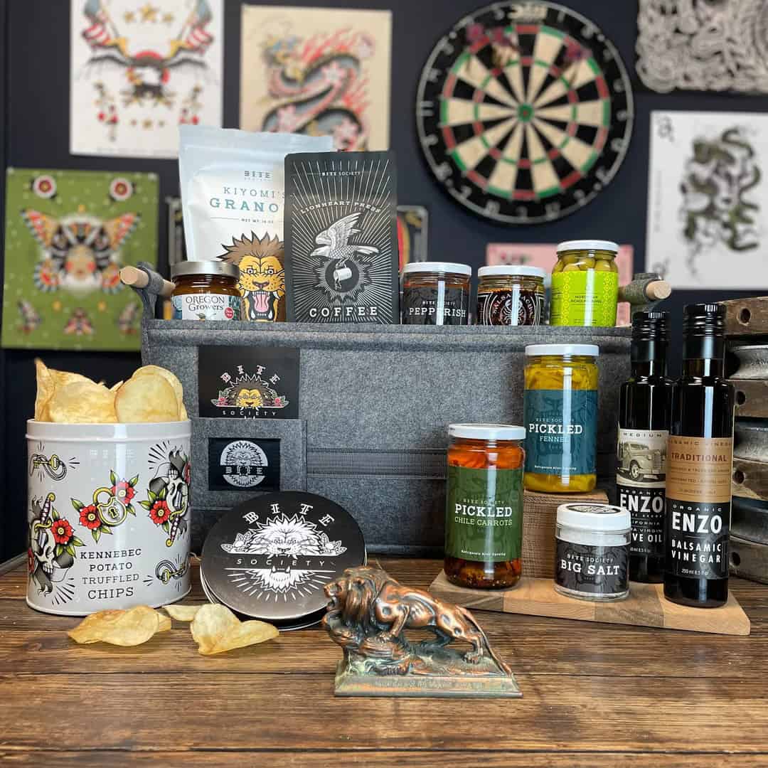 A gift basket on a wooden table features truffled potato chips, granola, coffee, pickled vegetables, salts, vinegars, and more—a perfect addition to any Holiday Gift Guide. Artistic wall hangings and a dartboard in the background add charm to this delightful scene.