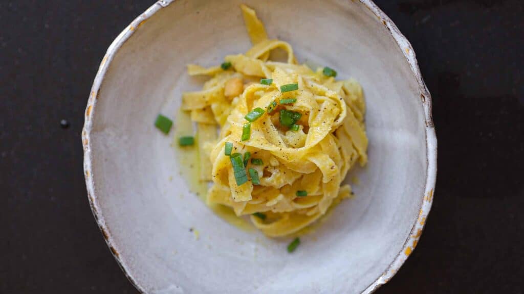 A bowl featuring chickpea pasta recipe topped with a creamy sauce and garnished with chopped green herbs, set against a dark background. The dish is served in a rustic, textured bowl.