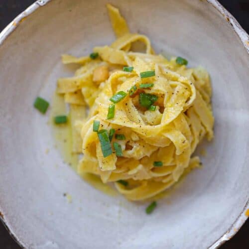 A bowl featuring chickpea pasta recipe topped with a creamy sauce and garnished with chopped green herbs, set against a dark background. The dish is served in a rustic, textured bowl.