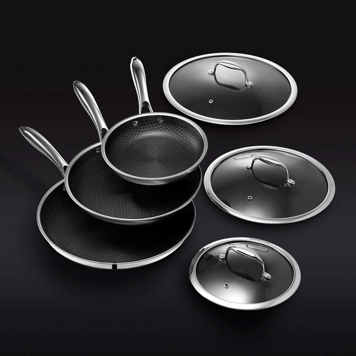 A set of three black frying pans with stainless steel handles, stacked and accompanied by three glass lids with metal handles, arranged on a black surface. Perfect for the Korean Vegan, this cookware makes an ideal addition to any 2024 Holiday Gift Guide.