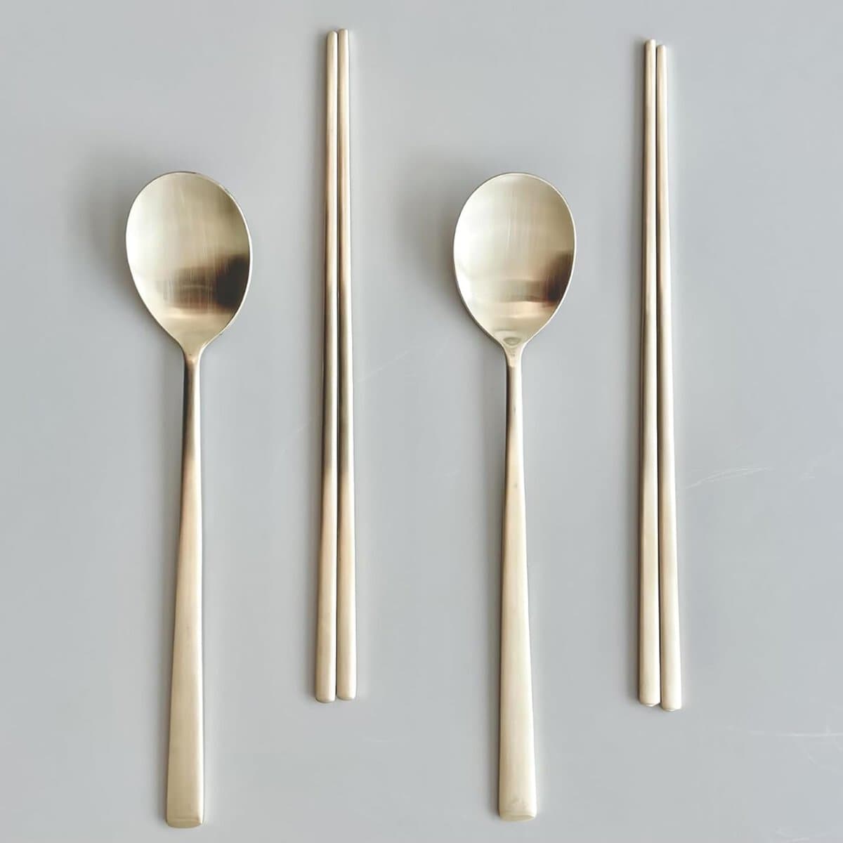 A neatly arranged set of two metal spoons paired with two pairs of metal chopsticks on a light grey background. The utensils have a sleek, modern design with a shiny finish.