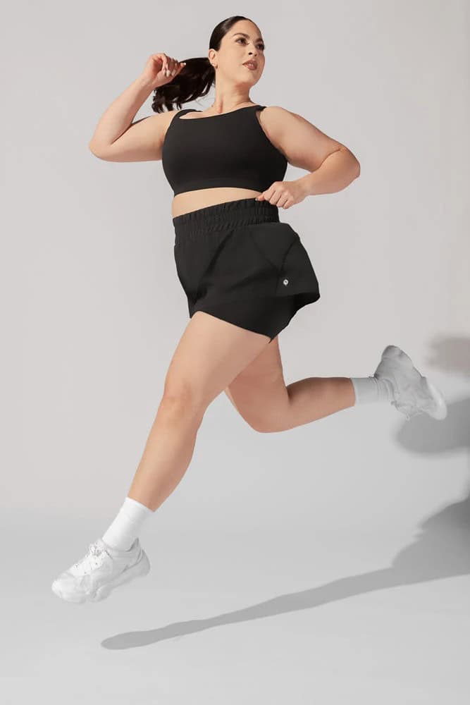 A person in a black sports bra and shorts jumps with energy and enthusiasm. They wear white sneakers and socks against a plain background, one arm bent and the other extended back.