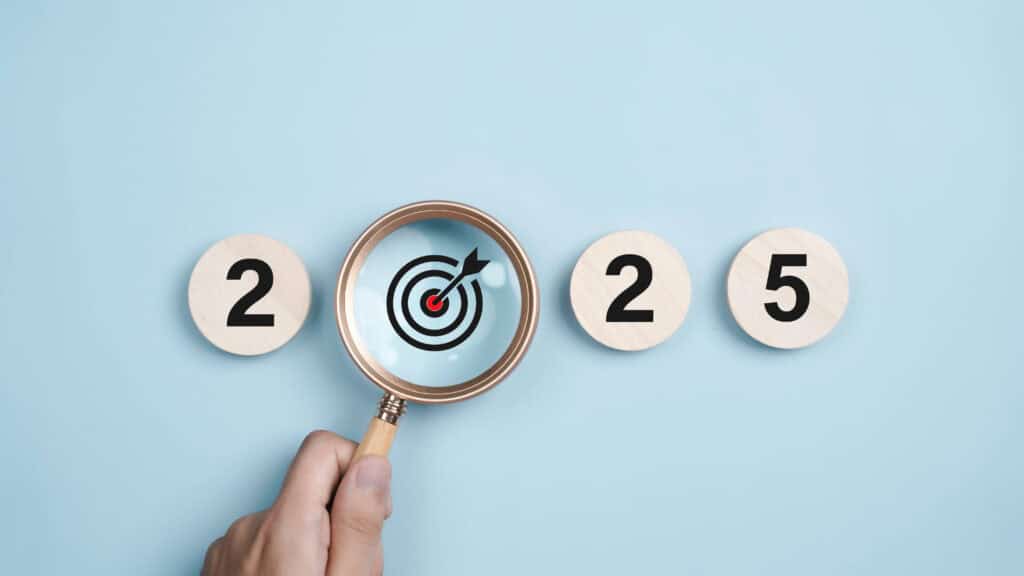 A hand with a magnifying glass zeroes in on a red and black target symbol nestled between the numbers 2025, embodying a New Years Resolution. Set against a light blue backdrop, the numbers rest on wooden circles, emphasizing focus and determination for the year ahead.