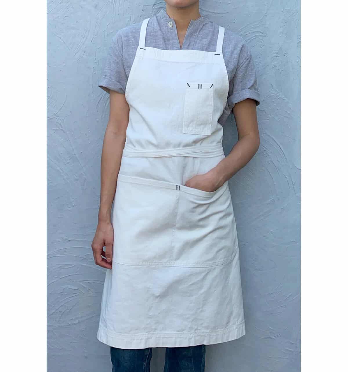 Standing against a textured gray wall, the person wears a short-sleeved gray shirt and a white apron with pockets. One hand rests thoughtfully in the apron pocket.