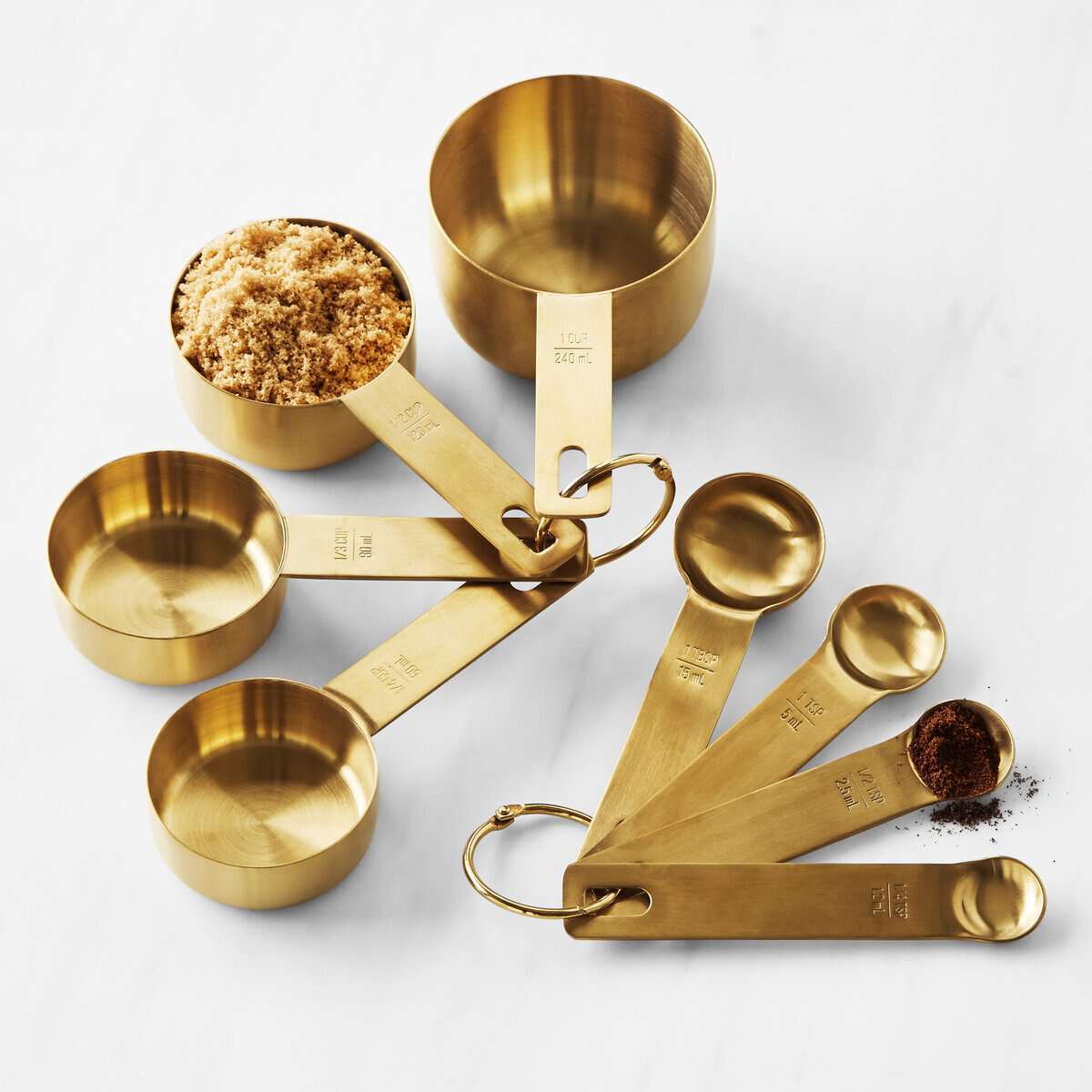 A perfect holiday gift, this set of gold measuring cups and spoons displayed on a white surface is ideal for any kitchen. Each cup filled with brown sugar and each spoon sprinkled with spices, offers precise measurement markings—an essential guide for any culinary adventure.