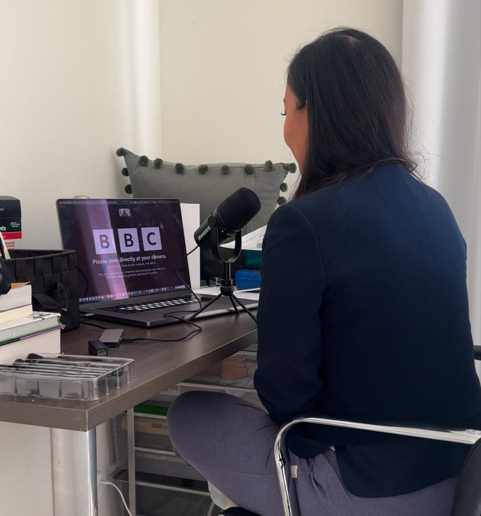 Joanne sits at a cluttered desk, surrounded by books and office supplies, speaking into a microphone on a video call with BBC displayed on her laptop.