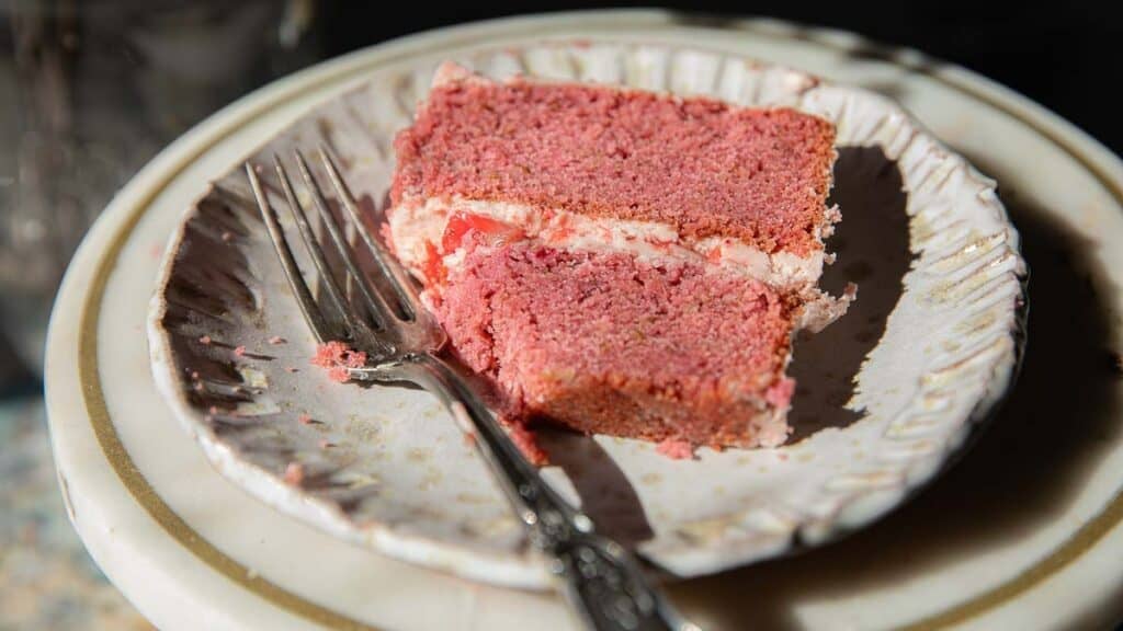 A slice of pink vegan strawberry cake with white frosting sits on a decorative plate with a fork beside it. The cake shows two layers, and bright lighting highlights its texture.