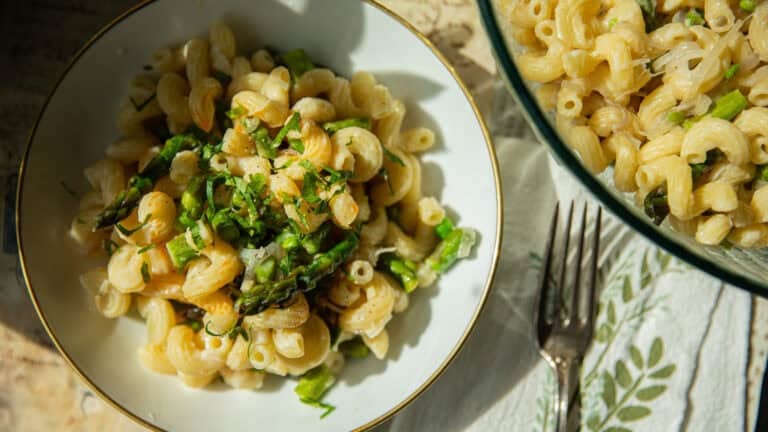 Recipes 19 A bowl of creamy asparagus pasta recipe with asparagus and herbs is served on a table. Another bowl of the same dish is nearby, accompanied by a fork resting on a floral-patterned cloth. Bright natural light highlights the foods textures and colors.