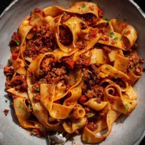 BEST Vegan Bolognese Recipe - Meaty and Delicious. 51 A bowl of wide, ribbon-like pasta topped with chunky vegan bolognese sauce, garnished with chopped herbs, and served on a gray plate.