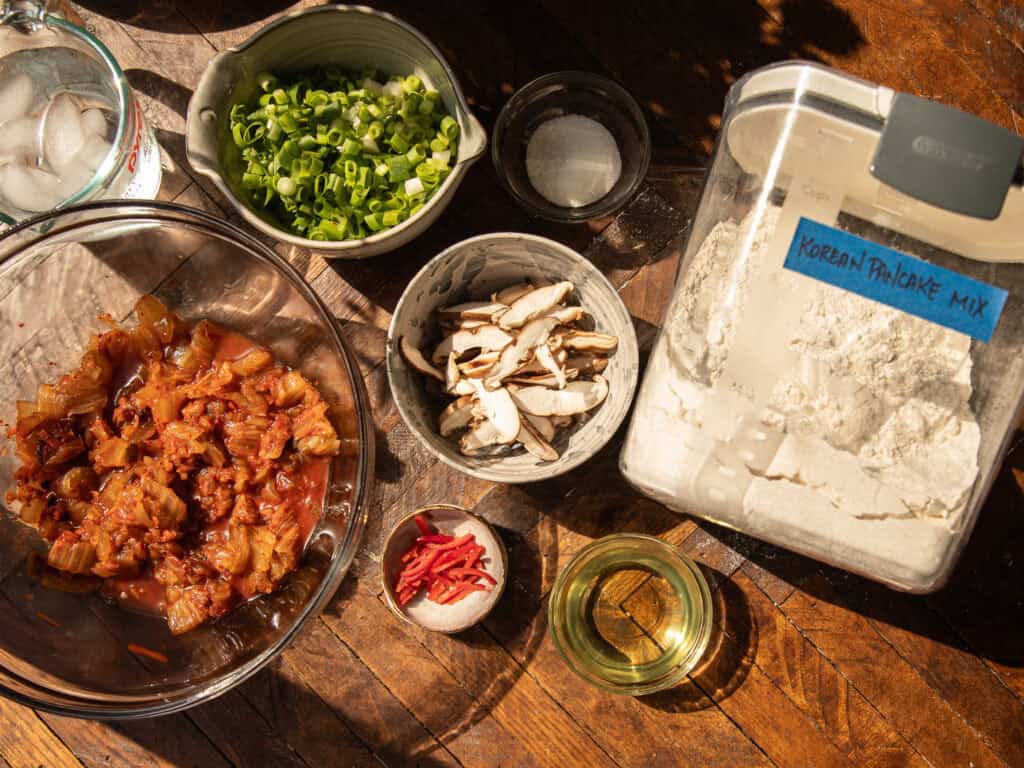 Easy and Crispy Korean Kimchi Pancakes (김치전) Recipe. 6 A top-down view of ingredients for Korean kimchi pancakes on a wooden table, including kimchi, sliced mushrooms, chopped green onions, pancake mix in a container, a bowl of ice water, oil, salt, and sliced red peppers.