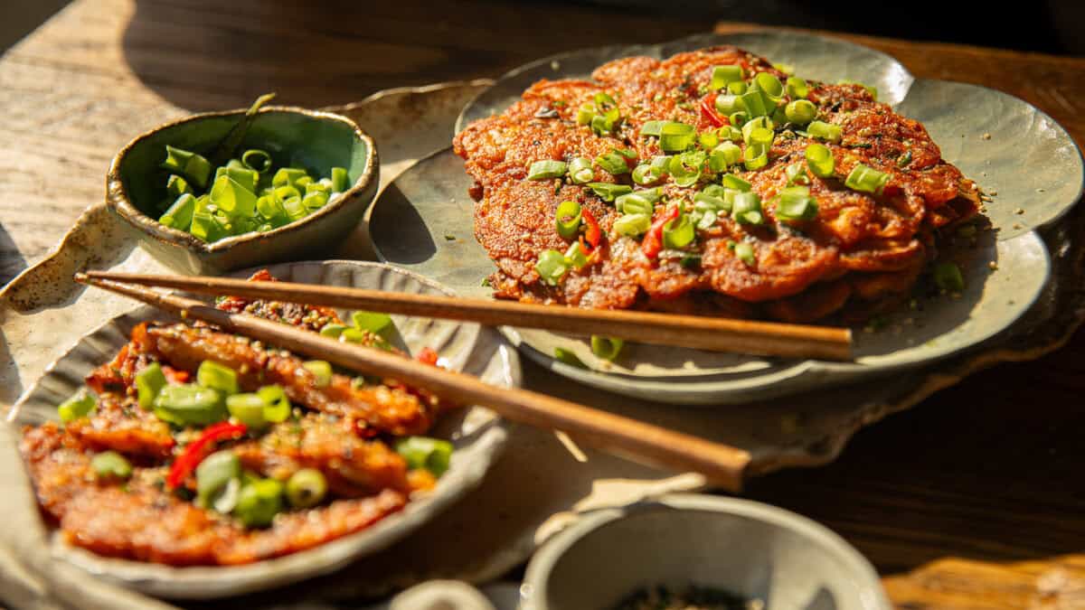 Plates of savory korean kimchi pancakes topped with chopped green onions are served with chopsticks and small bowls of extra green onions, all arranged on a wooden table in warm sunlight.