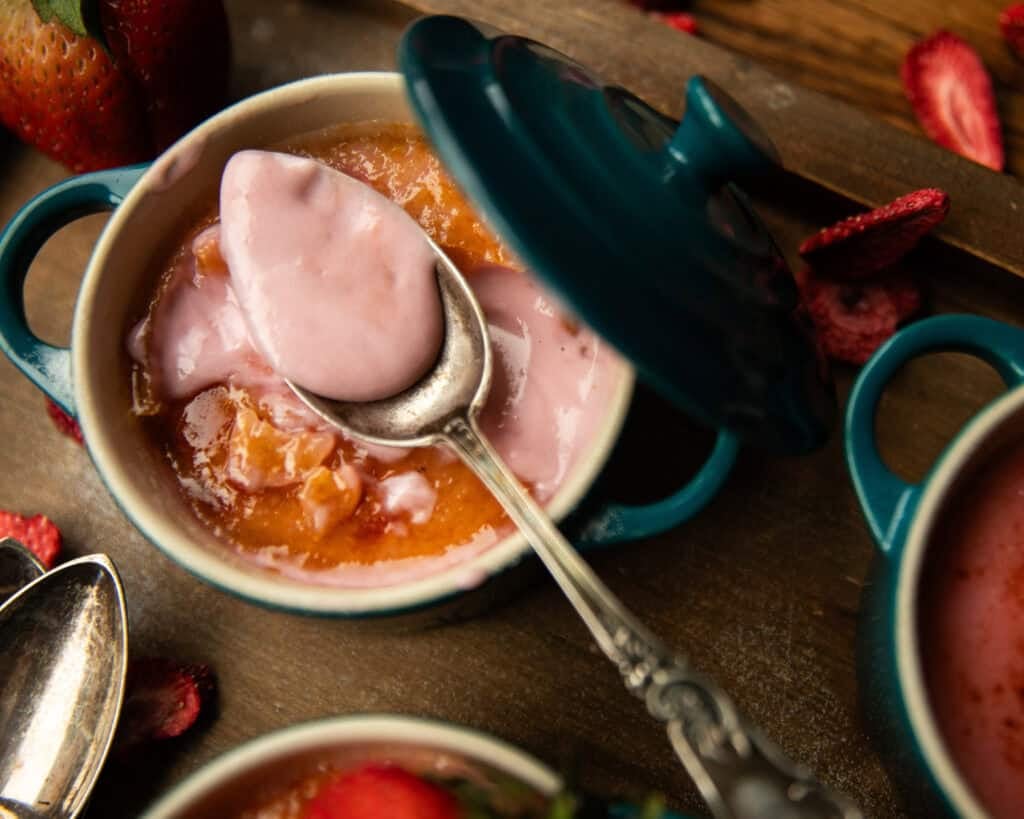 A small teal ceramic dish with a lid holds creamy pink Strawberry Crème Brûlée. A spoon scoops up some of the dessert, while fresh strawberries and another dish rest nearby on the wooden surface.