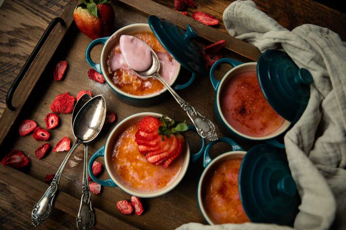 A wooden tray holds four small blue ramekins with strawberry crème brûlée, their caramelized tops adorned with fresh strawberries. Two vintage spoons rest nearby, and a light cloth is draped beside the dishes, with more sliced berries scattered around.