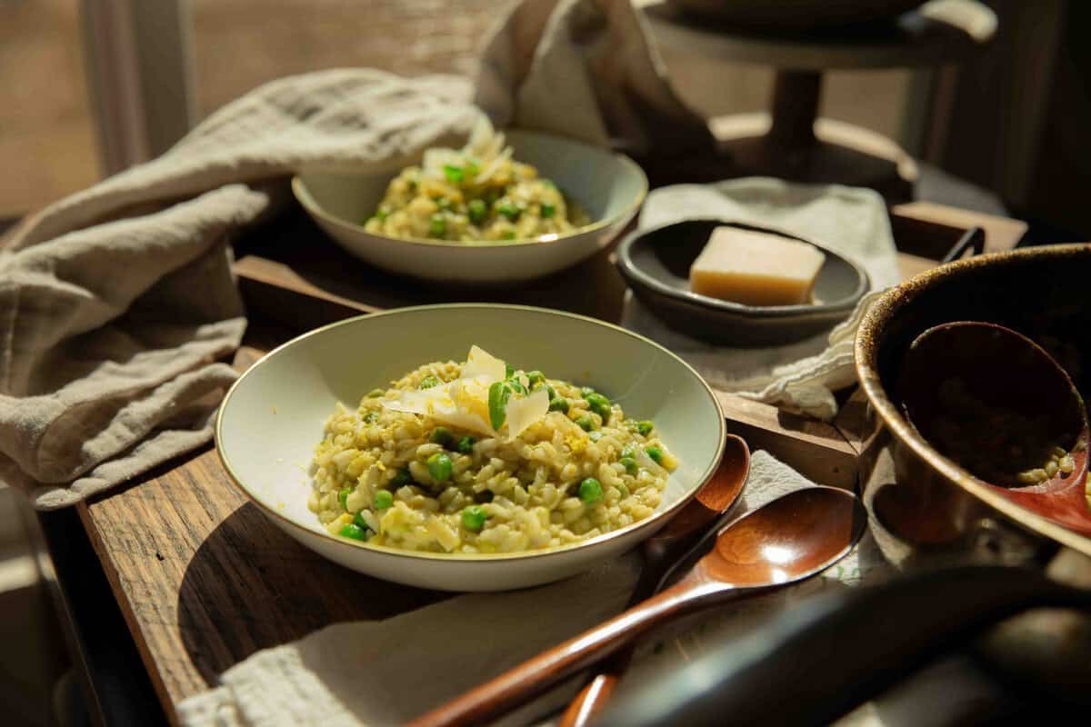 The BEST Sweet Green Pea Risotto Recipe (Vegan). 11 Two bowls of creamy green pea risotto with green peas and cheese sit on a tray, accompanied by a small plate with a wedge of cheese, wooden spoons, and a beige napkin in warm, natural sunlight.