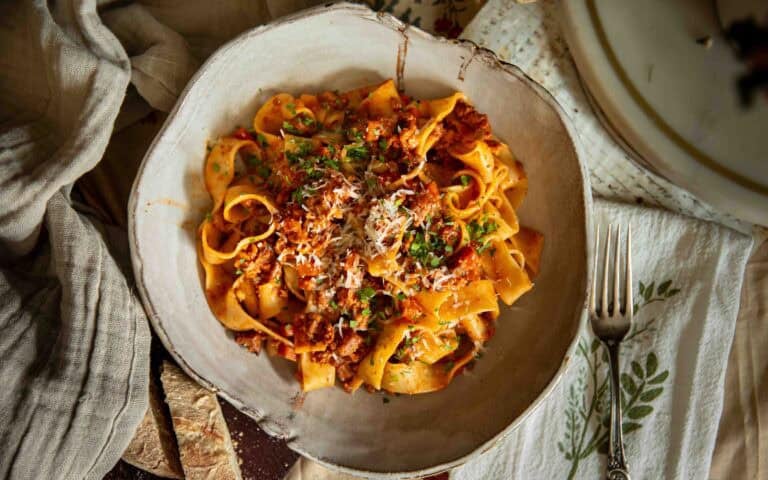 Recipes 14 A bowl of wide ribbon pasta topped with vegan bolognese, grated cheese, and herbs sits on a table with a fork, napkins, and slices of bread nearby.