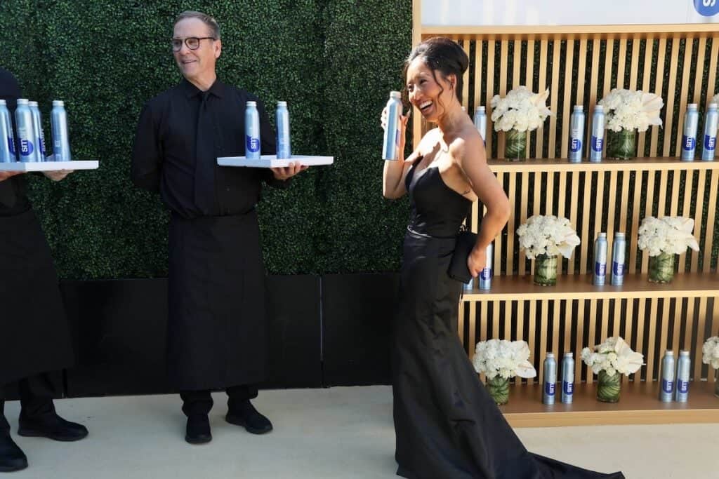 Mental toughness: "Shifting the line" of your distress tolerance. 3 Joanne, in a strapless black gown, smiles while holding a can, standing near a wooden shelf adorned with white flowers and cans. A server dressed in black stands beside her with a tray of cans; lush greenery forms the backdrop.