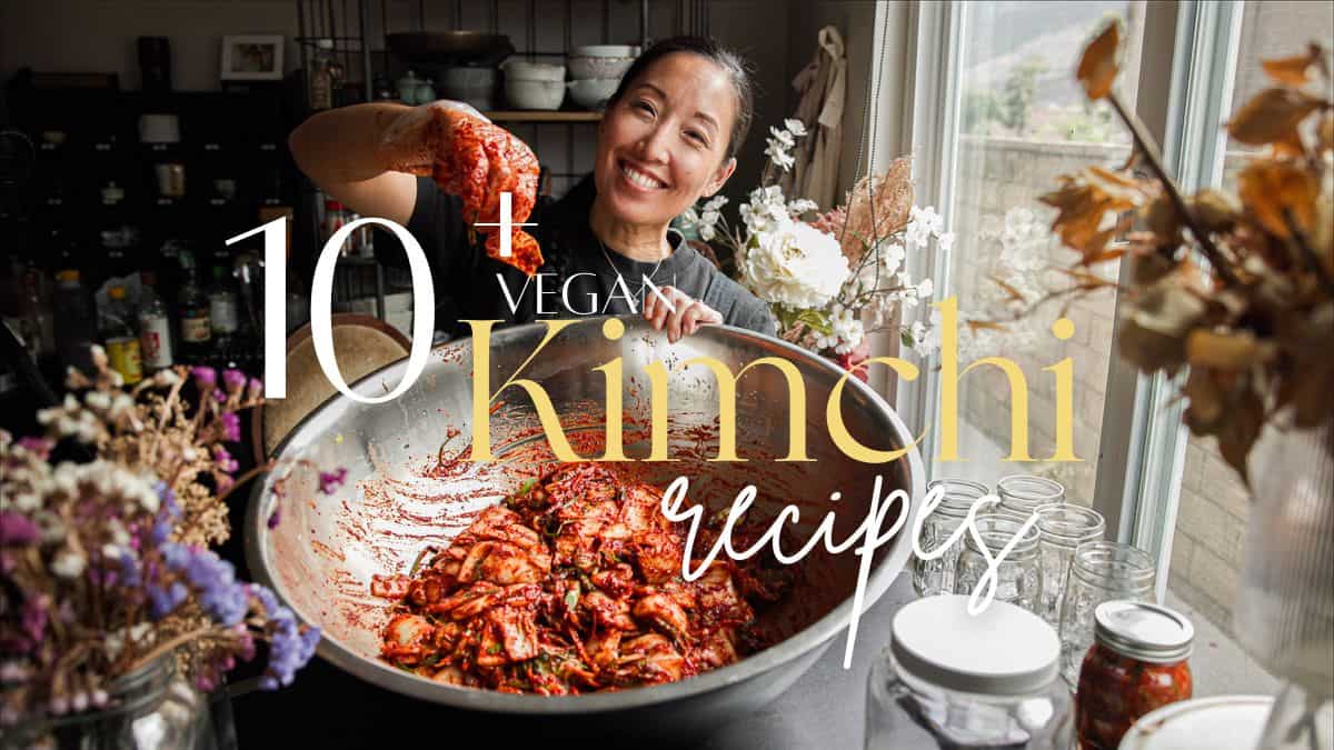 Easy and Crispy Korean Kimchi Pancakes (김치전) Recipe. 20 A woman smiles while mixing kimchi in a large metal bowl in a kitchen filled with jars and dried flowers. Overlaid text reads: 10+ Vegan Kimchi Recipes, including tangy kimchi pancakes.