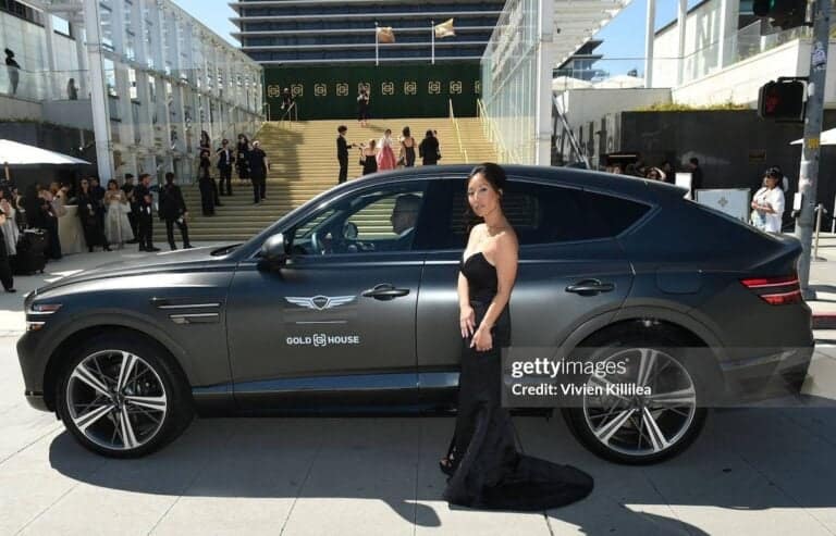A woman in a black gown stands beside a sleek black Genesis SUV with Gold House branding, exuding mental toughness as she poses confidently before a grand staircase and building, while people gather in the background.